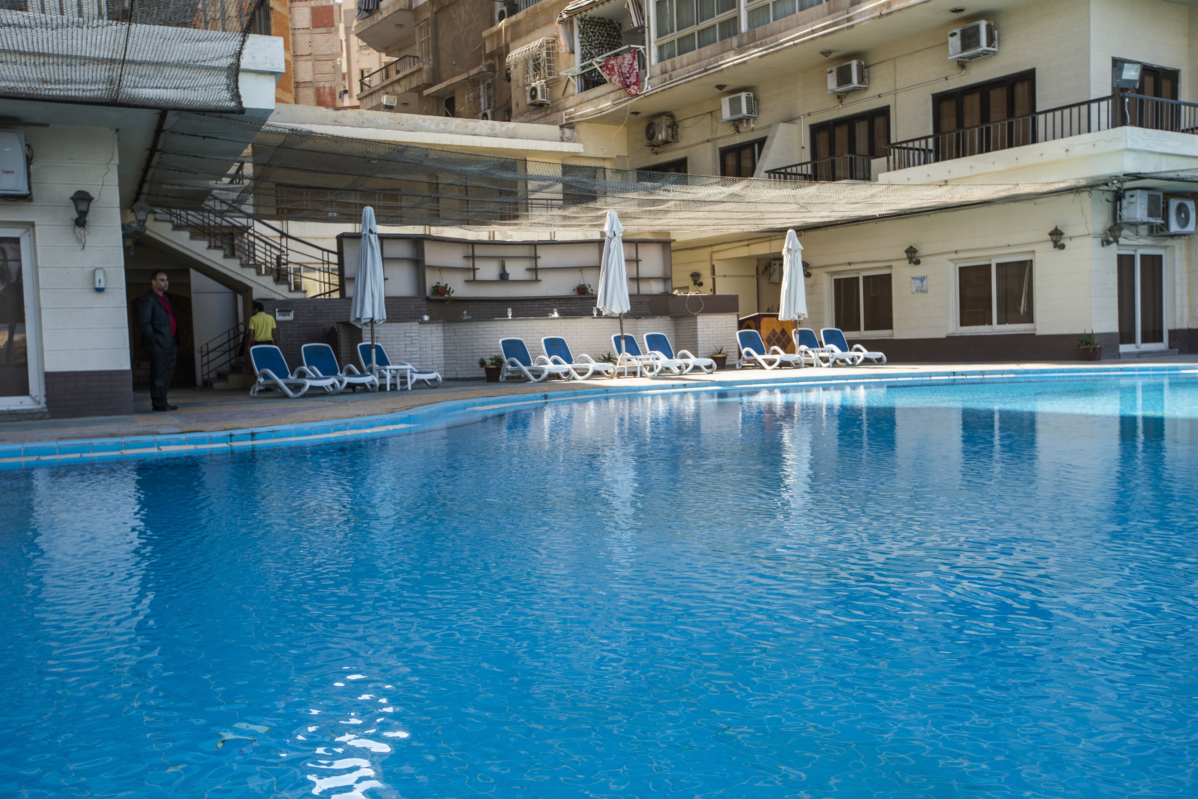 A swimming pool with lounge chairs and umbrellas, surrounded by a building with multiple balconies and air conditioning units.