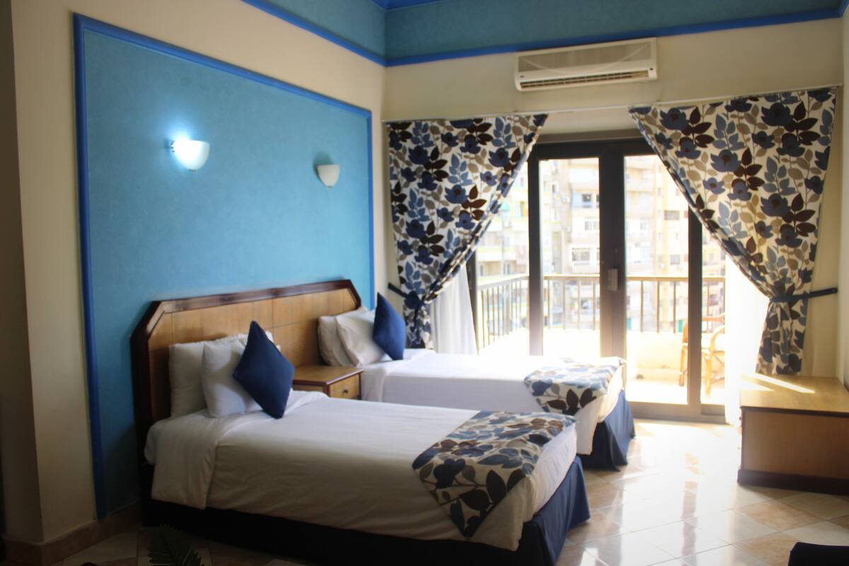 A hotel room with two beds, a window with curtains, a wall-mounted air conditioner, and a view of the outside balcony.