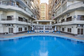 A swimming pool surrounded by balconies with air conditioning units, a central seating area with lounge chairs, and a view of a high-rise building in the background. - Aifu Resort (Alexandria)