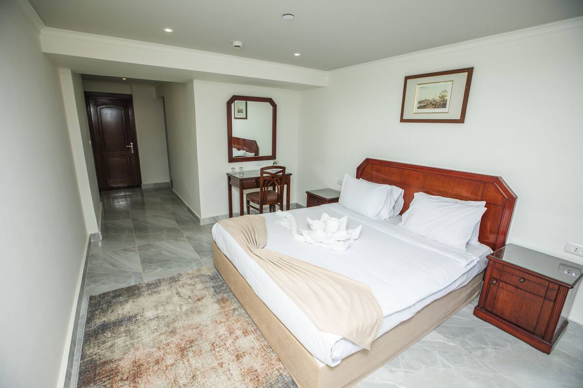 A hotel room with a large bed, a wooden nightstand, a desk with a chair, a mirror, and a framed picture on the wall.
