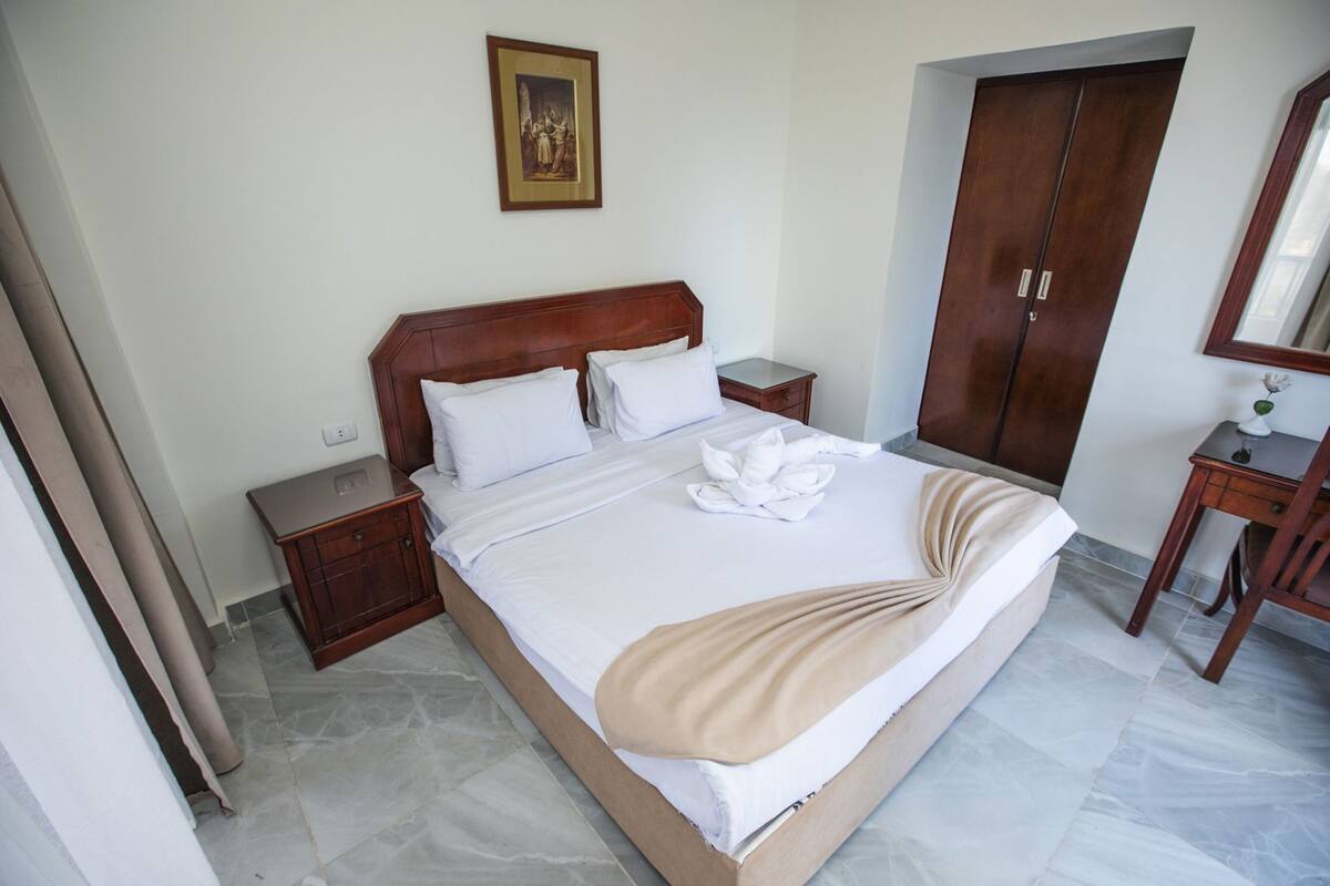 A hotel room with a large bed, wooden headboard, a nightstand, a wardrobe, and a small desk with a mirror.