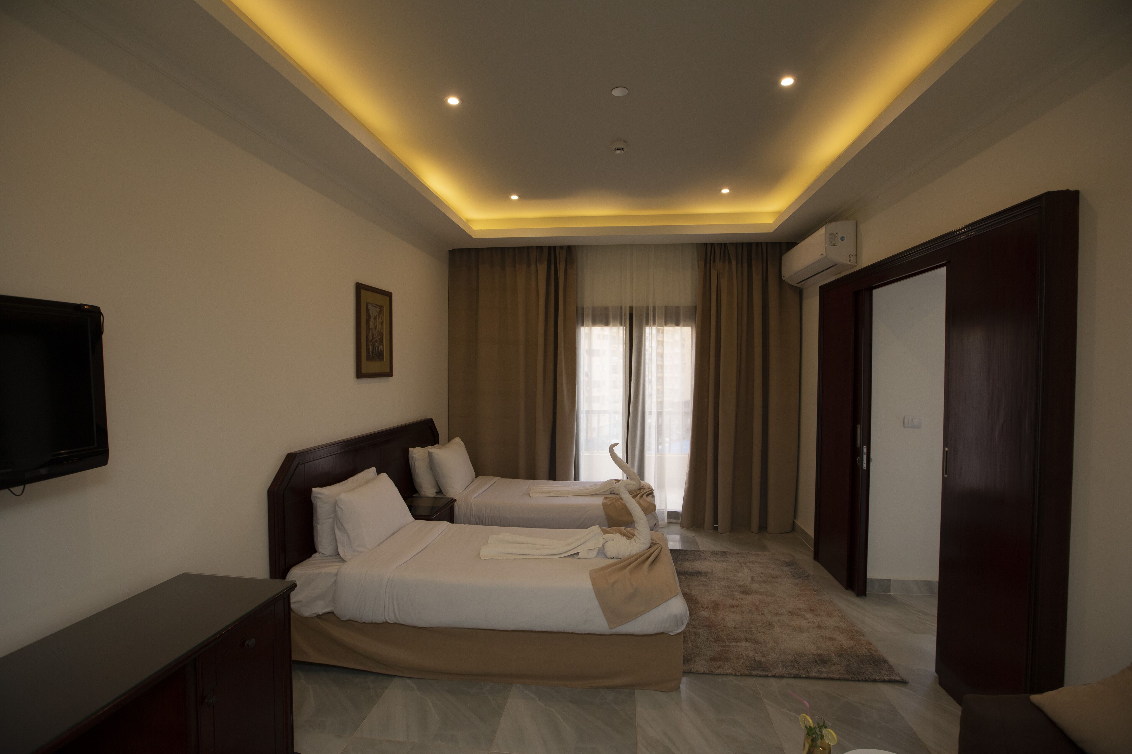 A hotel room with two beds, a flat-screen TV, a wardrobe, and a large window with curtains.