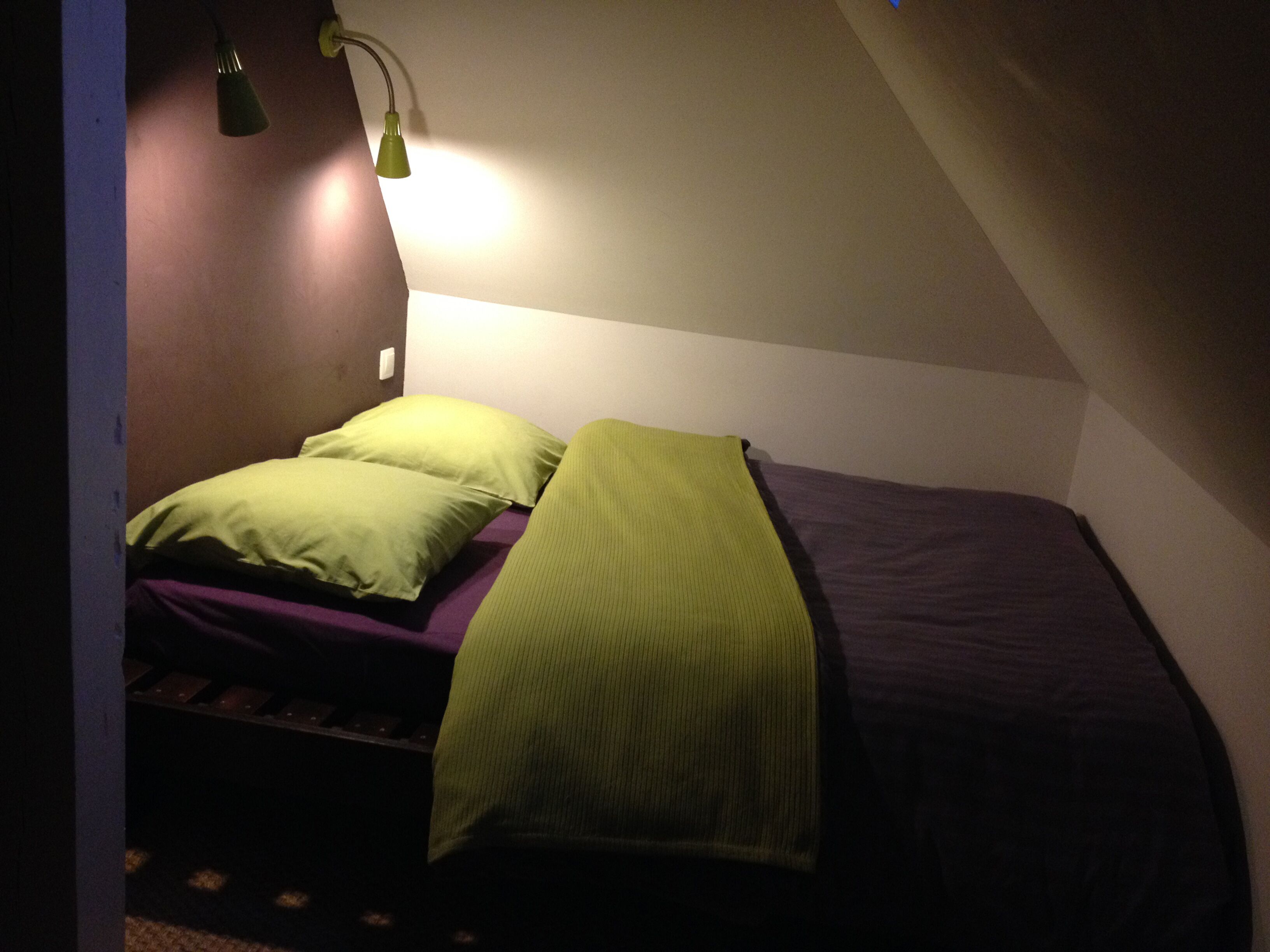 Ein kleiner, schwach beleuchteter Raum mit einem Bett, einem grünen Kopfkissen und einer grünen Decke.
