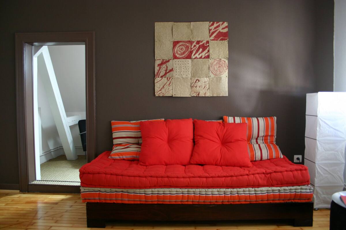 Une chambre avec un lit rouge, des oreillers à rayures et une courtepointe accrochée au mur.