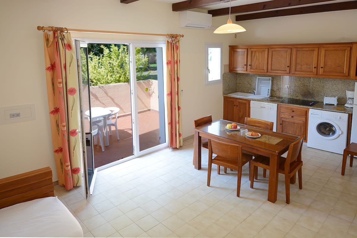 Une pièce comprenant un lit, une table à manger avec des chaises, un coin cuisine avec des meubles en bois, un lave-linge et une porte-fenêtre coulissante donnant sur un espace extérieur.