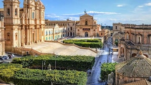 A historic plaza with classical architecture, including a prominent church with a dome and bell towers, surrounded by well-maintained greenery and a clear sky. - B&B del Carmine (Noto)