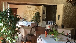 A rustic dining area with stone walls, wooden beams, and metal chairs. Tables are set with white tablecloths and potted plants. - La Mandarine (Piolenc)