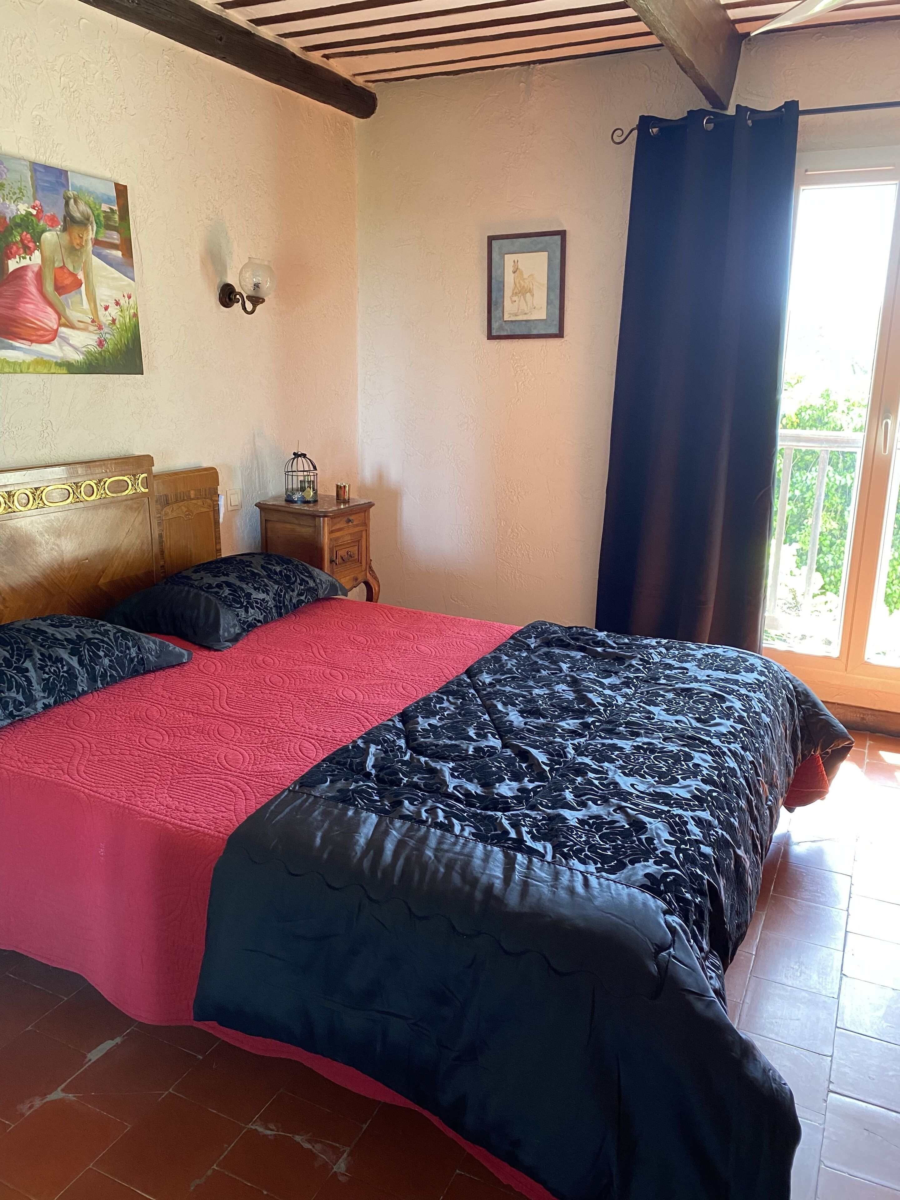 Een slaapkamer met een tweepersoonsbed, een klein nachtkastje, een wandklok en een raam met gordijnen.