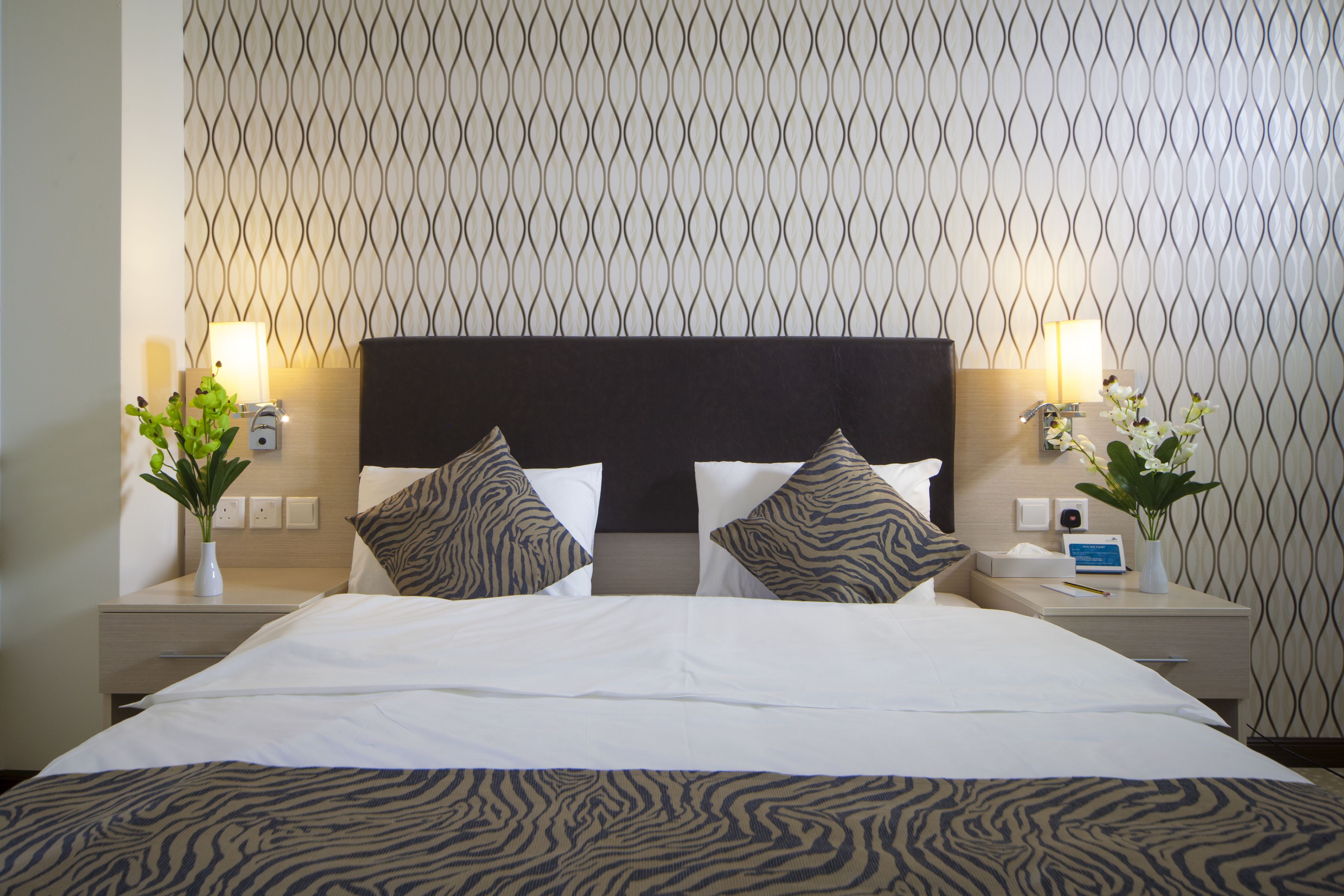 Ein ordentlich bezogenes Bett mit einer zebra-gemusterten Tagesdecke, zwei passende Kissen und ein dunkles Kopfteil. Zwei Nachttische mit Lampen, eine kleine Vase mit Blumen und eine Uhr sind ebenfalls vorhanden.