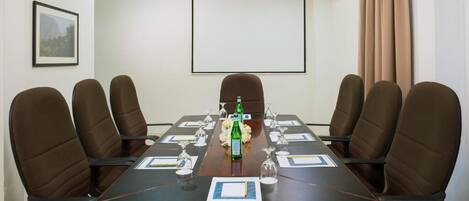 Un salón de conferencias con una larga mesa, seis sillas, una pantalla de proyector y una botella de agua.