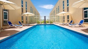 Une piscine rectangulaire bordée de chaises longues et d’un parasol de chaque côté, entourée d’immeubles beiges dotés de grandes fenêtres.
