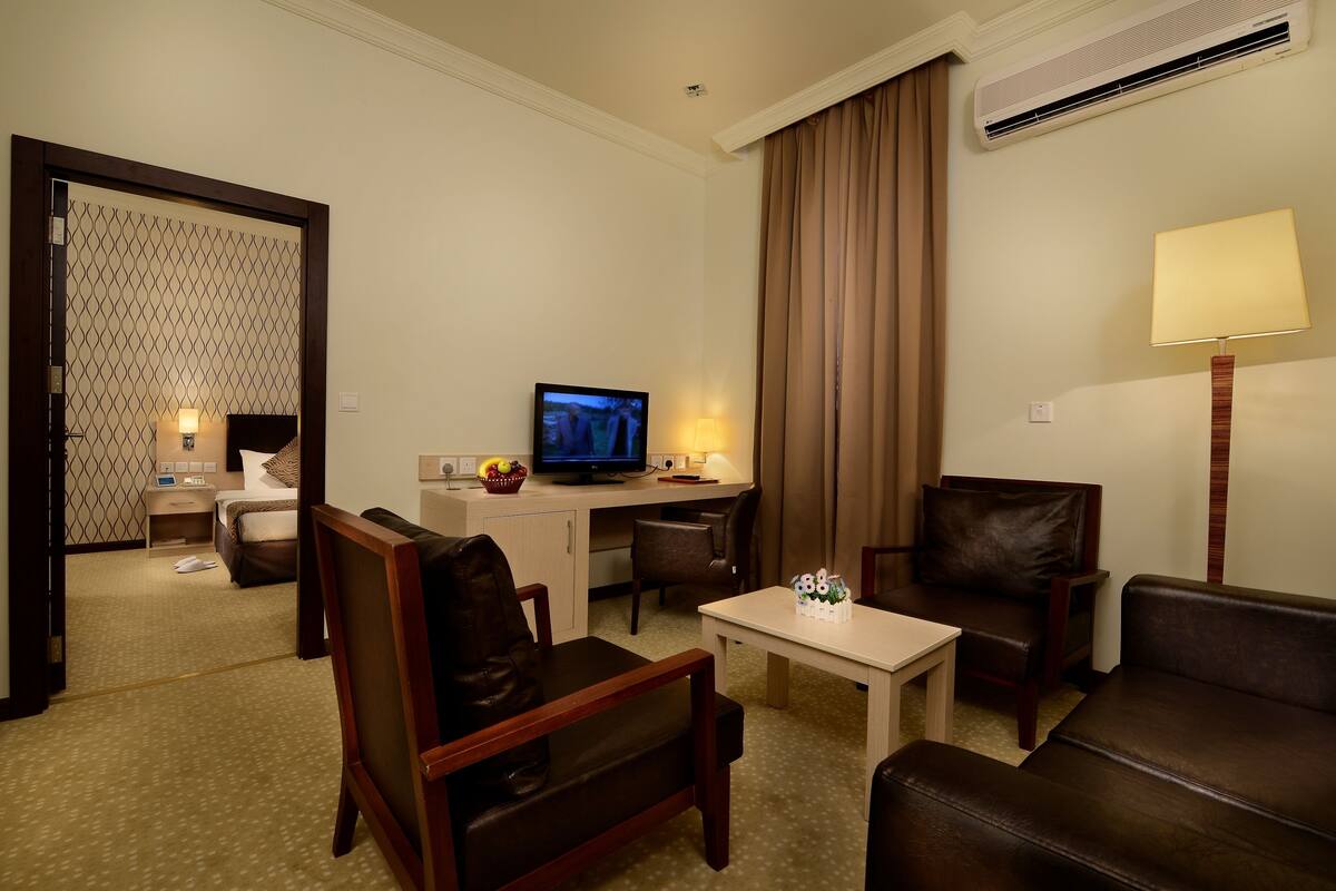 Habitación de hotel con cama, escritorio con computadora, televisor, mesa de centro y dos sillones de cuero.