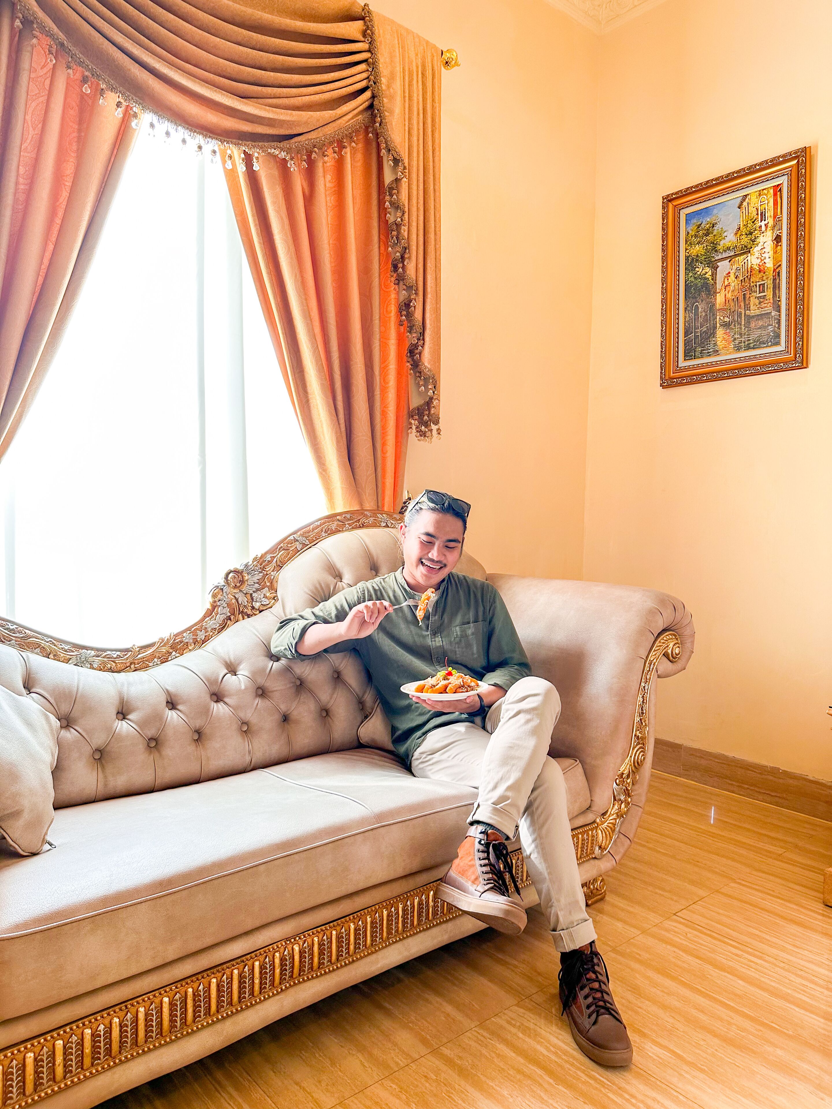 A person sitting on a tufted sofa with ornate detailing, holding a plate of food.