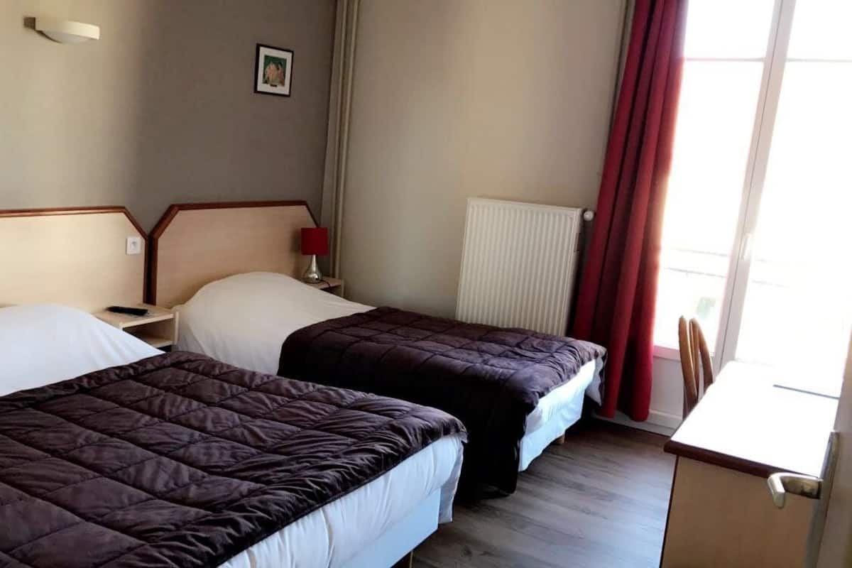 Ein Hotelzimmer mit zwei Betten, einem Nachttisch mit Lampe, einem Bild an der Wand und einem Fenster mit Vorhängen.