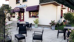 An outdoor seating area with wicker chairs and tables, a building with a sign reading "Le Saint-Rémy," and potted plants. - Hotel Le Saint Remy (Saint-Remy)