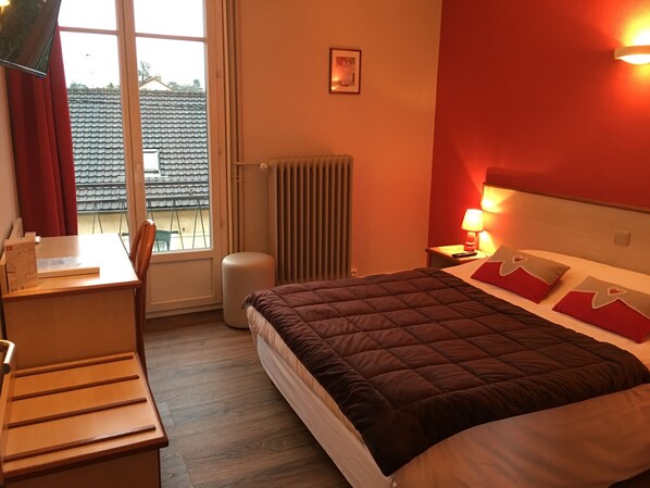 Habitación de hotel con cama, escritorio, silla, radiador y ventana con cortinas.