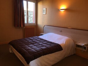 Una cama bien hecha con un edredón marrón, una mesita de noche con una lámpara y una ventana con cortinas.