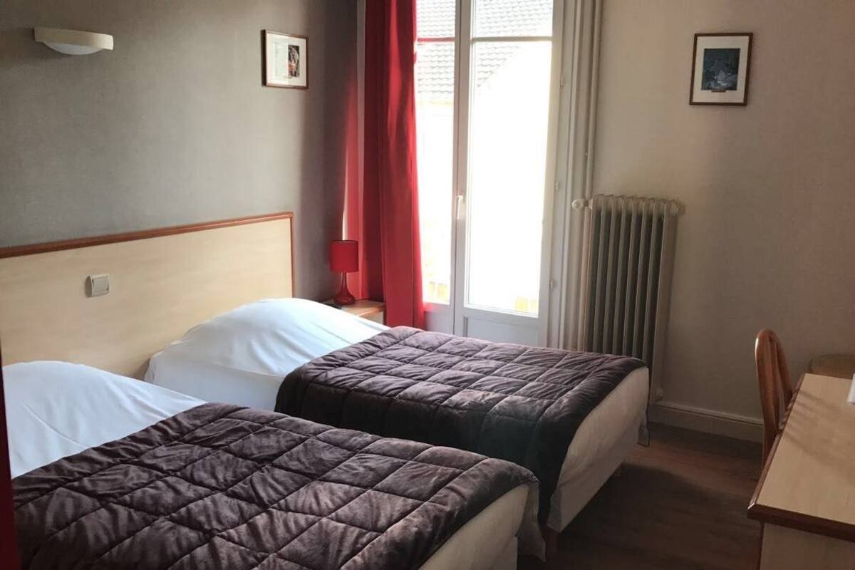Ein Hotelzimmer mit zwei Betten, einem Tisch und einem Stuhl. Es gibt ein Fenster mit Vorhängen, eine Heizung und gerahmte Bilder an der Wand.