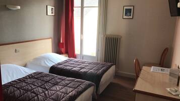 A hotel room with two beds, a table, and a chair. There is a window with curtains, a radiator, and framed pictures on the wall.