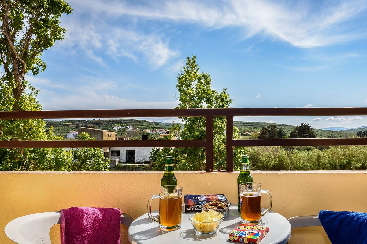 A balcony with a table set for two, featuring a bottle of beer, a glass of beer, a bowl of chips, and a pack of UNO cards.