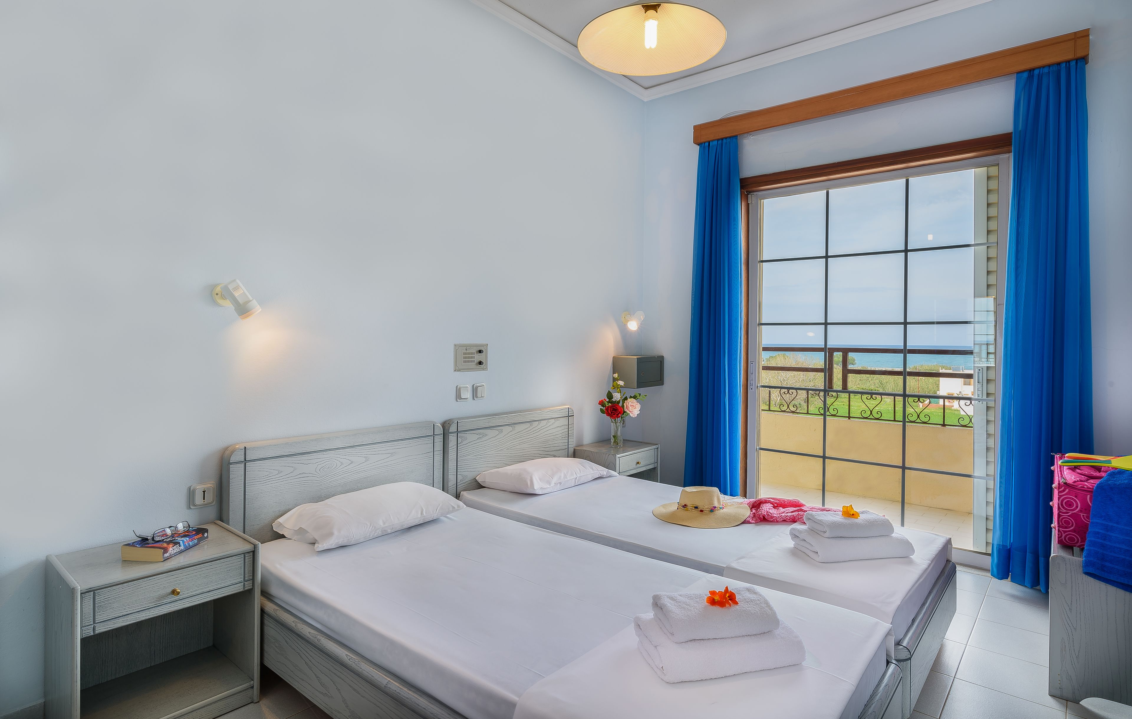 A hotel room with two beds, a window with blue curtains, a nightstand with a lamp, and a view of the outdoors.