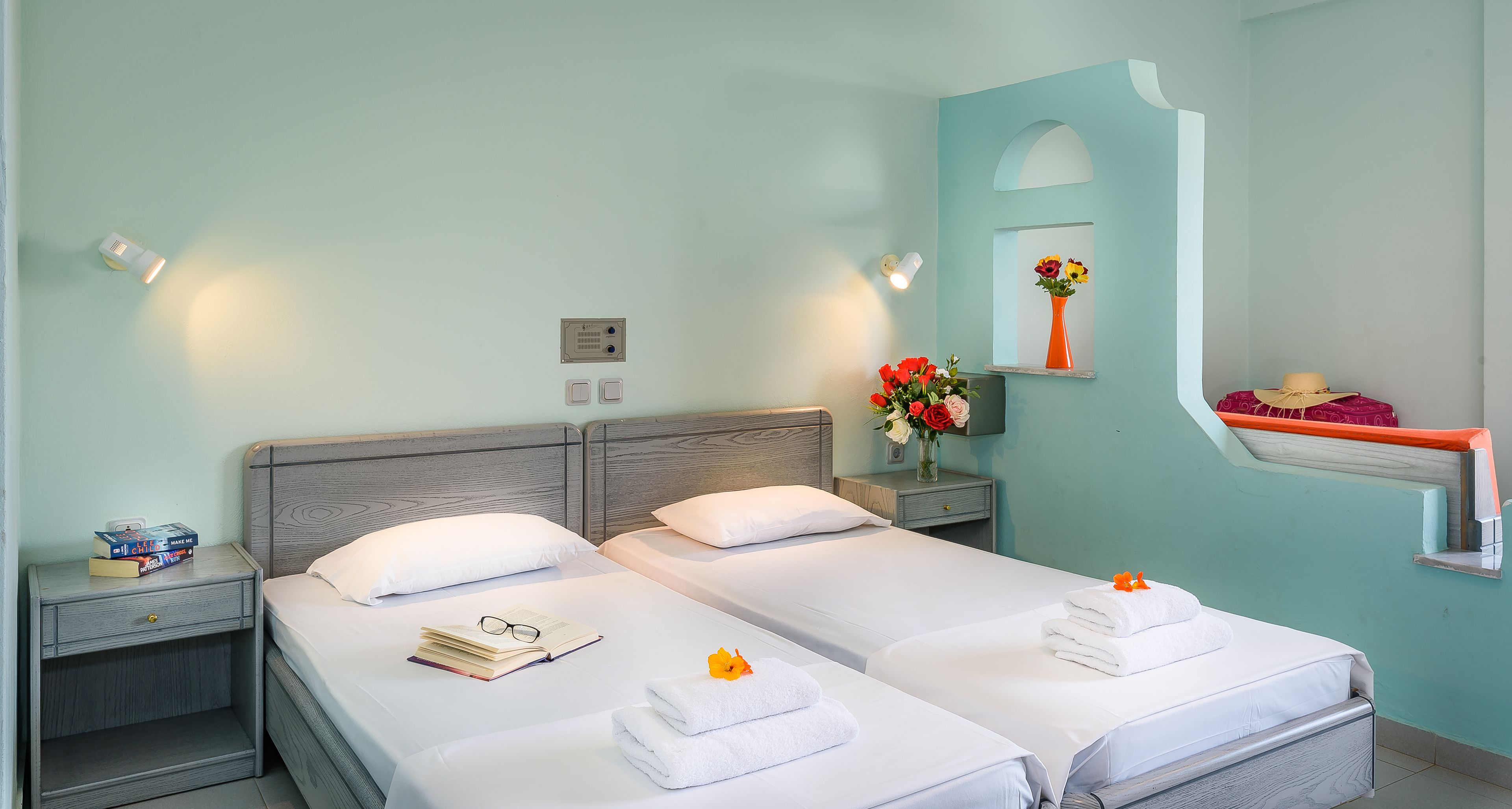 A hotel room with two beds, a nightstand with books and glasses, a vase with flowers, and a mirror with a shelf.