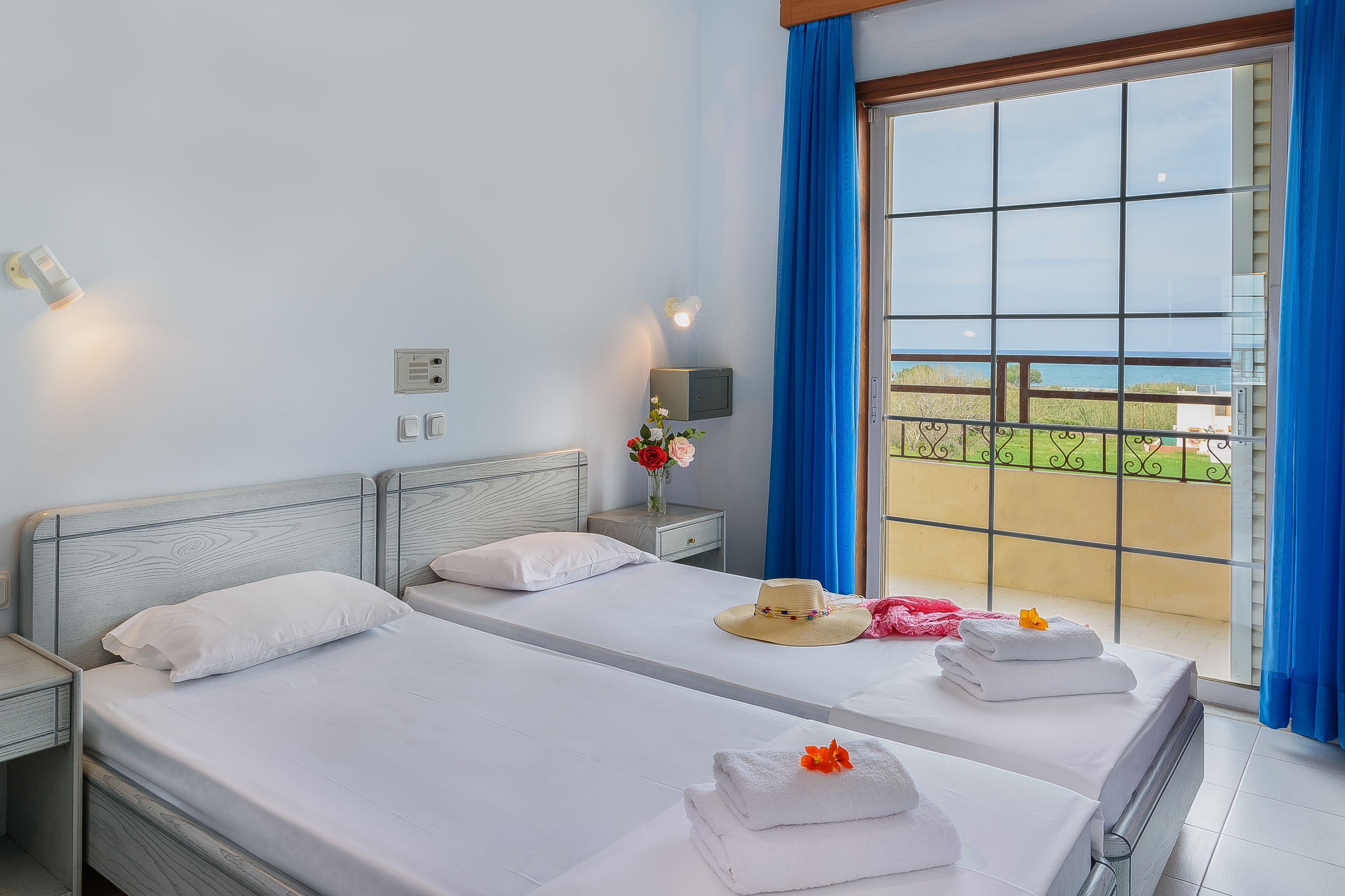 Habitación de hotel con dos camas, una mesita de noche, una pequeña mesa con un florero y vistas al exterior a través de un ventanal con cortinas azules.