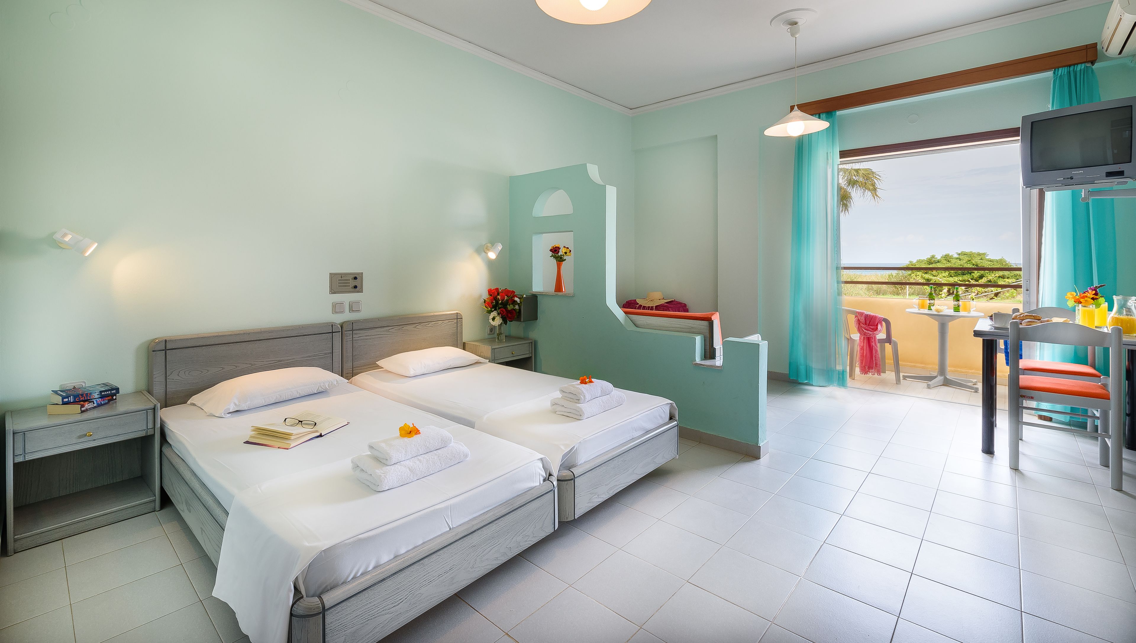 A hotel room with two beds, a small table with a chair, a TV, and a balcony with a view.