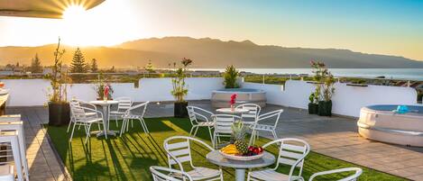 Una terrazza sul tetto con sedie e tavoli in metallo bianco, un piccolo tavolo con frutta, e vista sul mare e sulle montagne sullo sfondo.