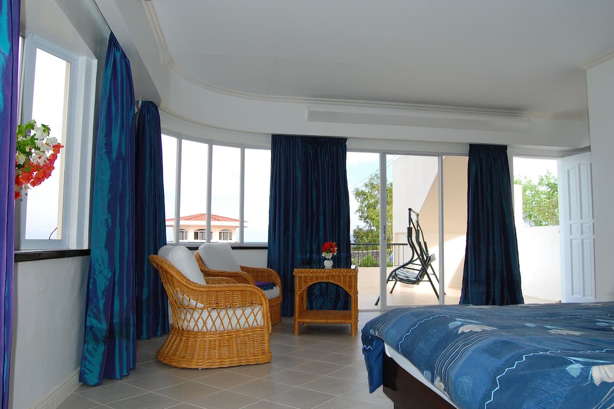 A bedroom with a bed, wicker chairs, a small table with a vase, and large windows with curtains.