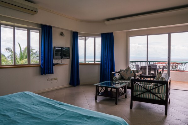 A hotel room with a bed, a glass table, a sofa, and a TV. The room has large windows with curtains and a view of the sea. - Sherwood Bay Resort & Aqua Sports (Dauis)
