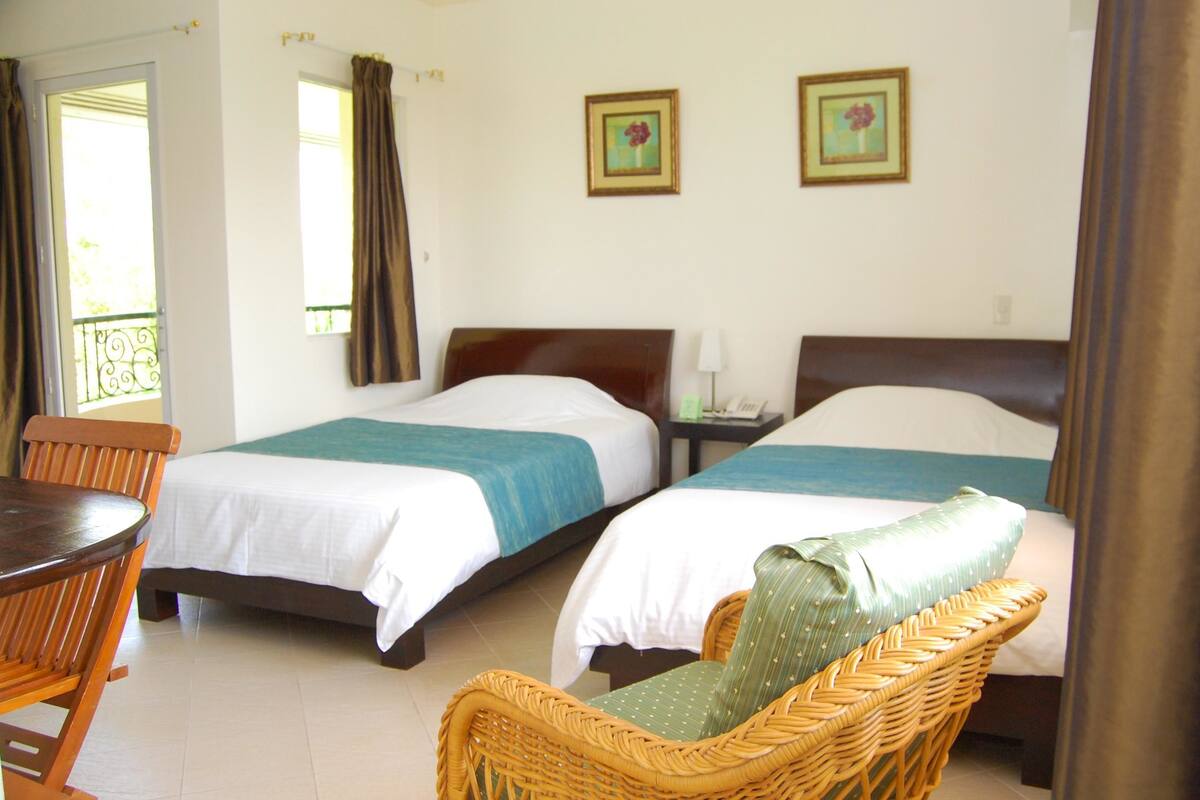 A hotel room with two beds, a wicker chair, a small table, and two framed pictures on the wall.