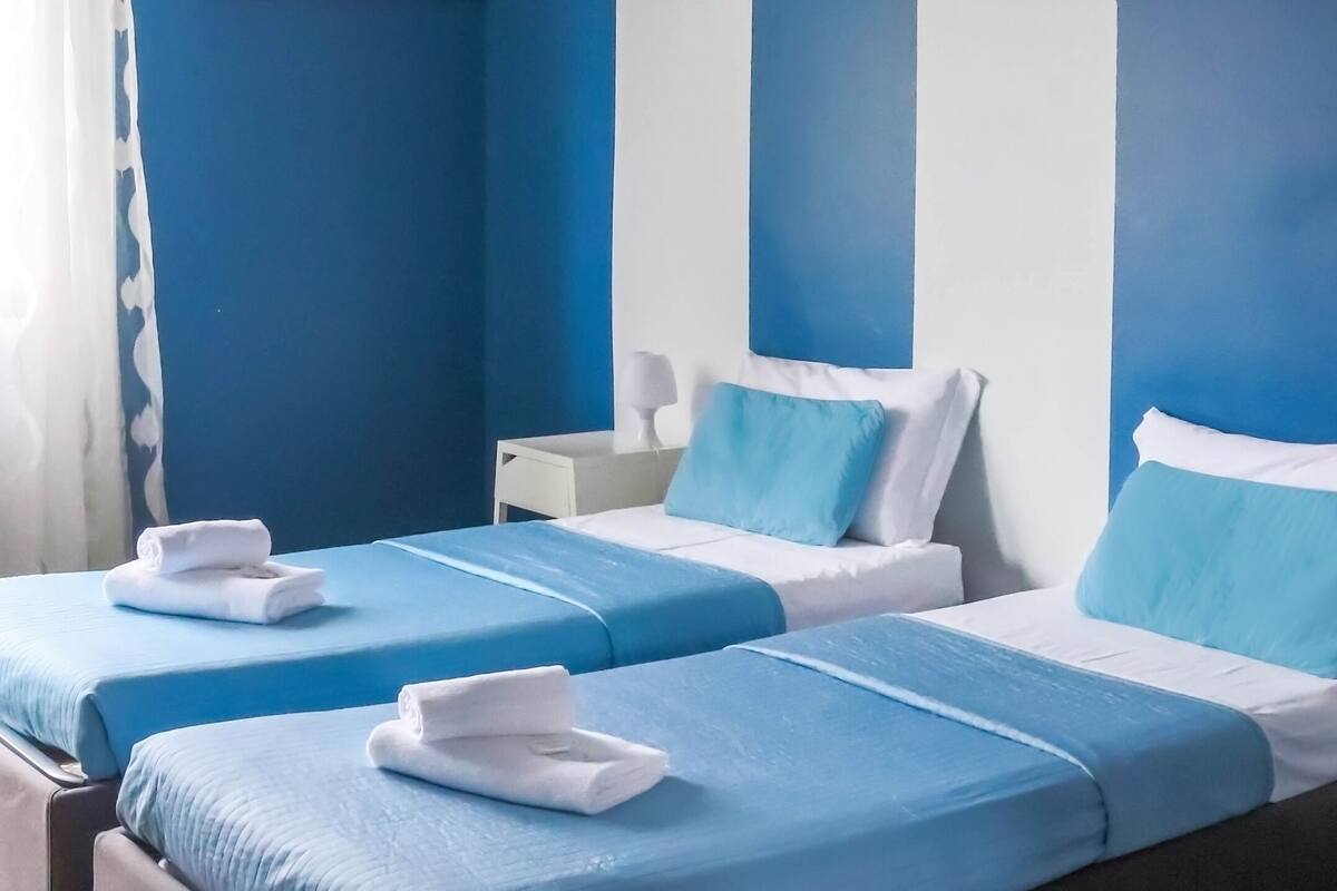 Een hotelkamer met twee bedden, elk met blauw beddengoed en witte kussens, een schilderij aan de muur en een witte lamp op een nachtkastje.