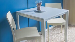 Een kamer met een witte tafel en stoelen, een wandtelevisie en een glazen vaas met bloemen op de tafel.