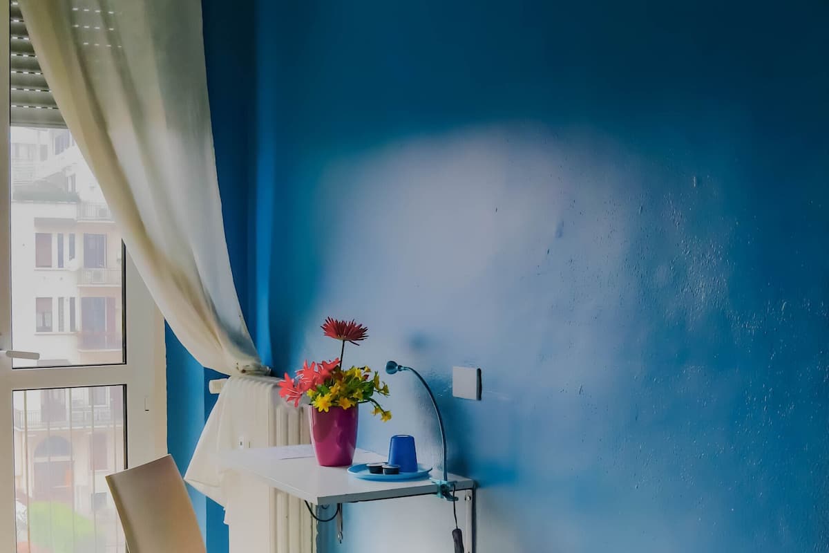 Een slaapkamer met een bed, een stoel, een plank met bloemen en een raam met gordijnen.