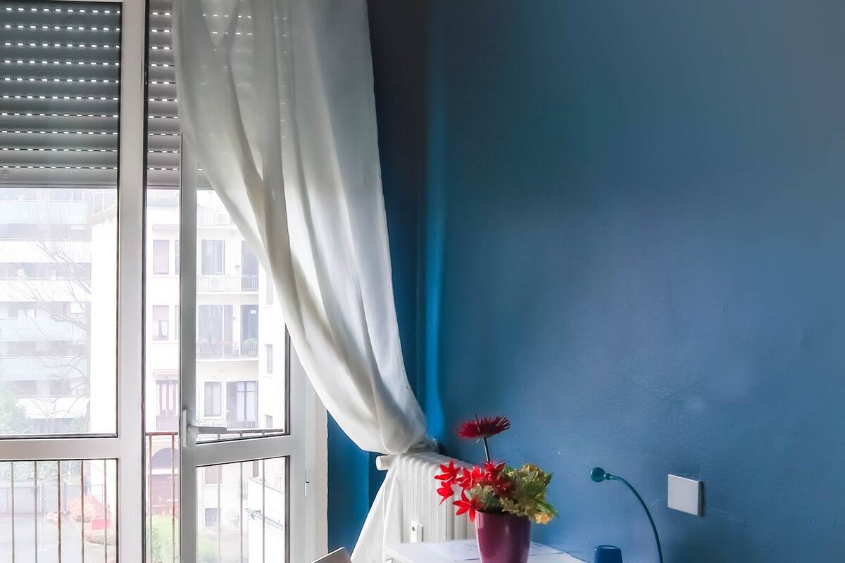 Een slaapkamer met een bed, een stoel, een bureau met lamp, een potplant en uitzicht op buiten.