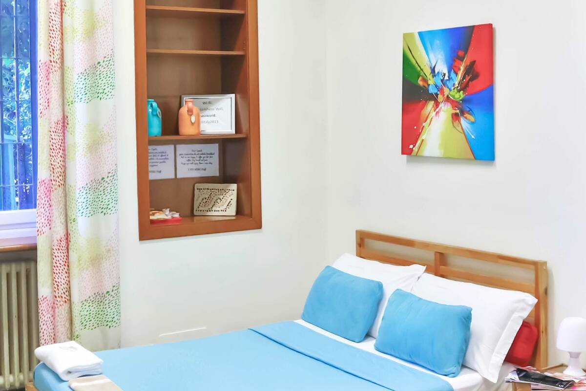 Een kamer met een houten bed, blauw beddengoed, een plank met spullen, een kleurrijk schilderij en een raam met gordijn.