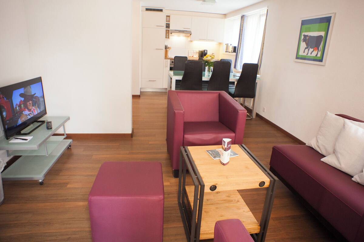A living room with a TV, a sofa, a coffee table, and a dining area in the background.
