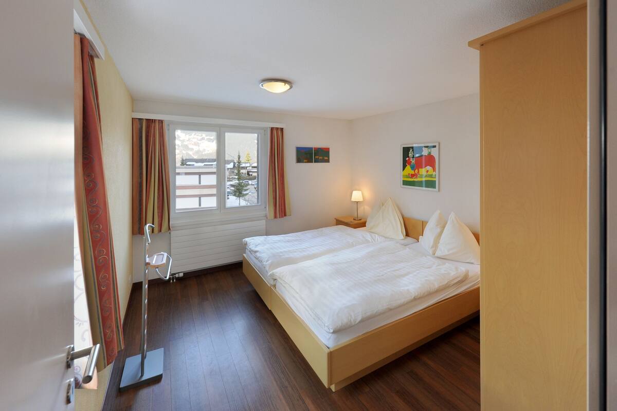 A hotel room with a large bed, a nightstand with a lamp, a window with curtains, and a painting on the wall.