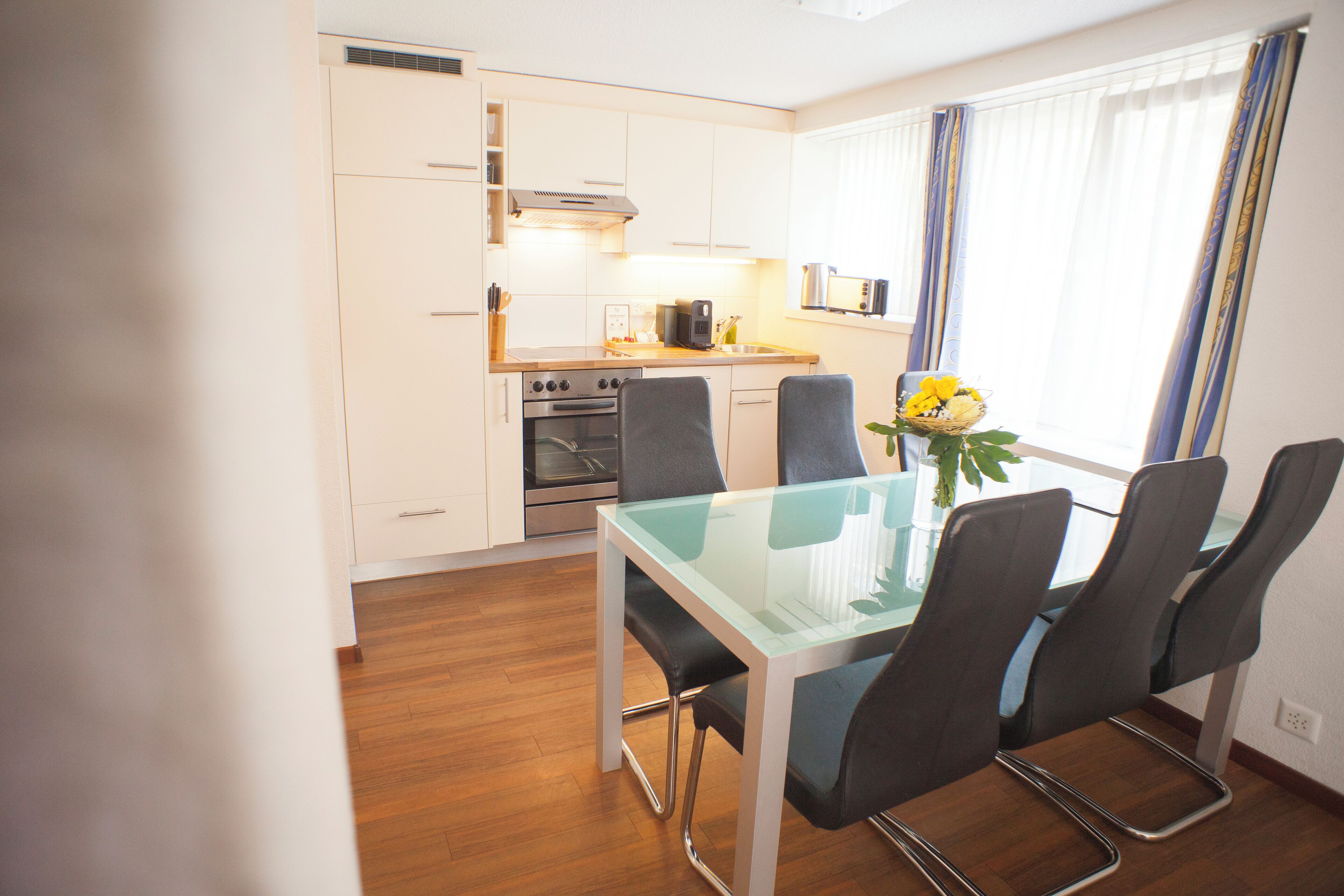 A modern kitchen with white cabinets, stainless steel appliances, and a glass dining table with four chairs.