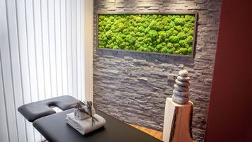 Un cuarto de spa con pared de piedra, panel de pared cubierto de musgo verde, una camilla de masaje y una escultura de madera.