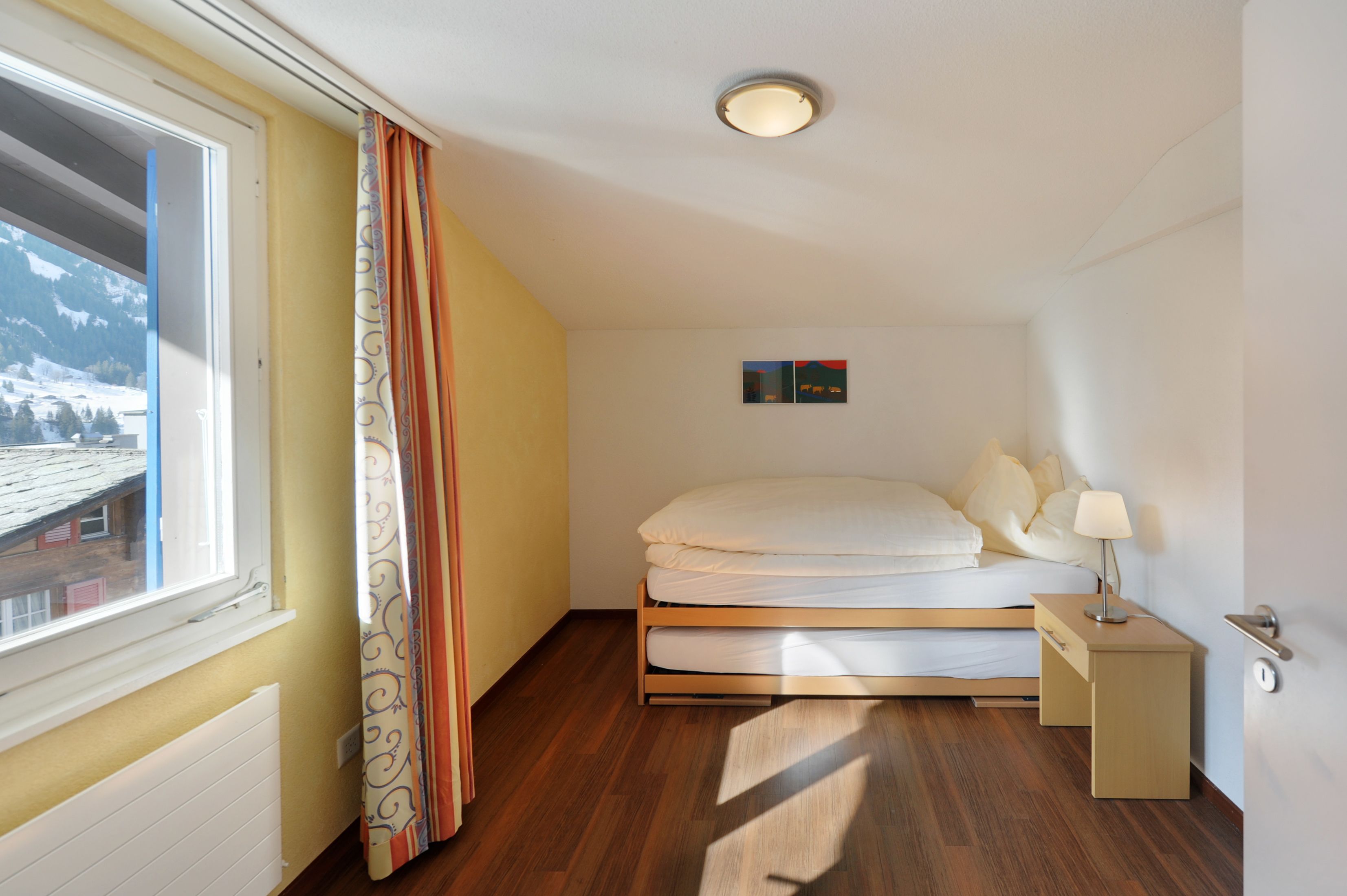 A bedroom with a bed, a nightstand, a window with curtains, a radiator, and a small painting on the wall.