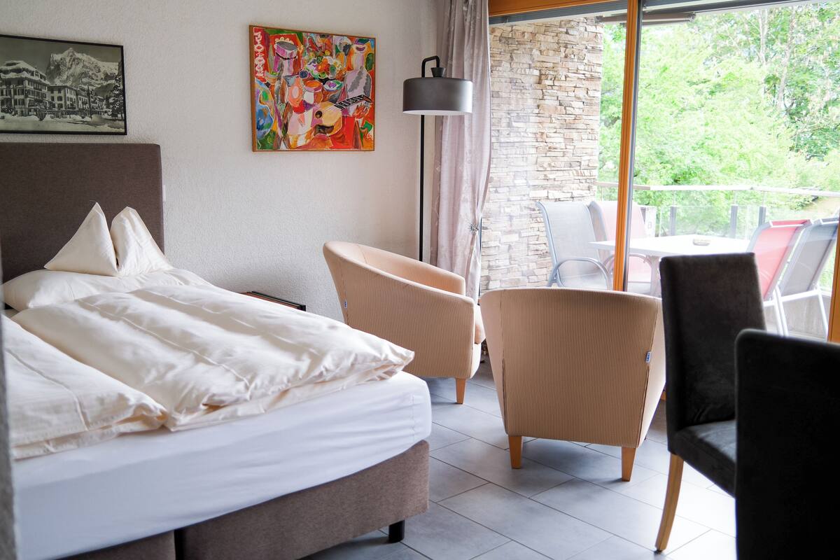 A hotel room with a bed, a chair, a table, and a balcony with a view of trees.