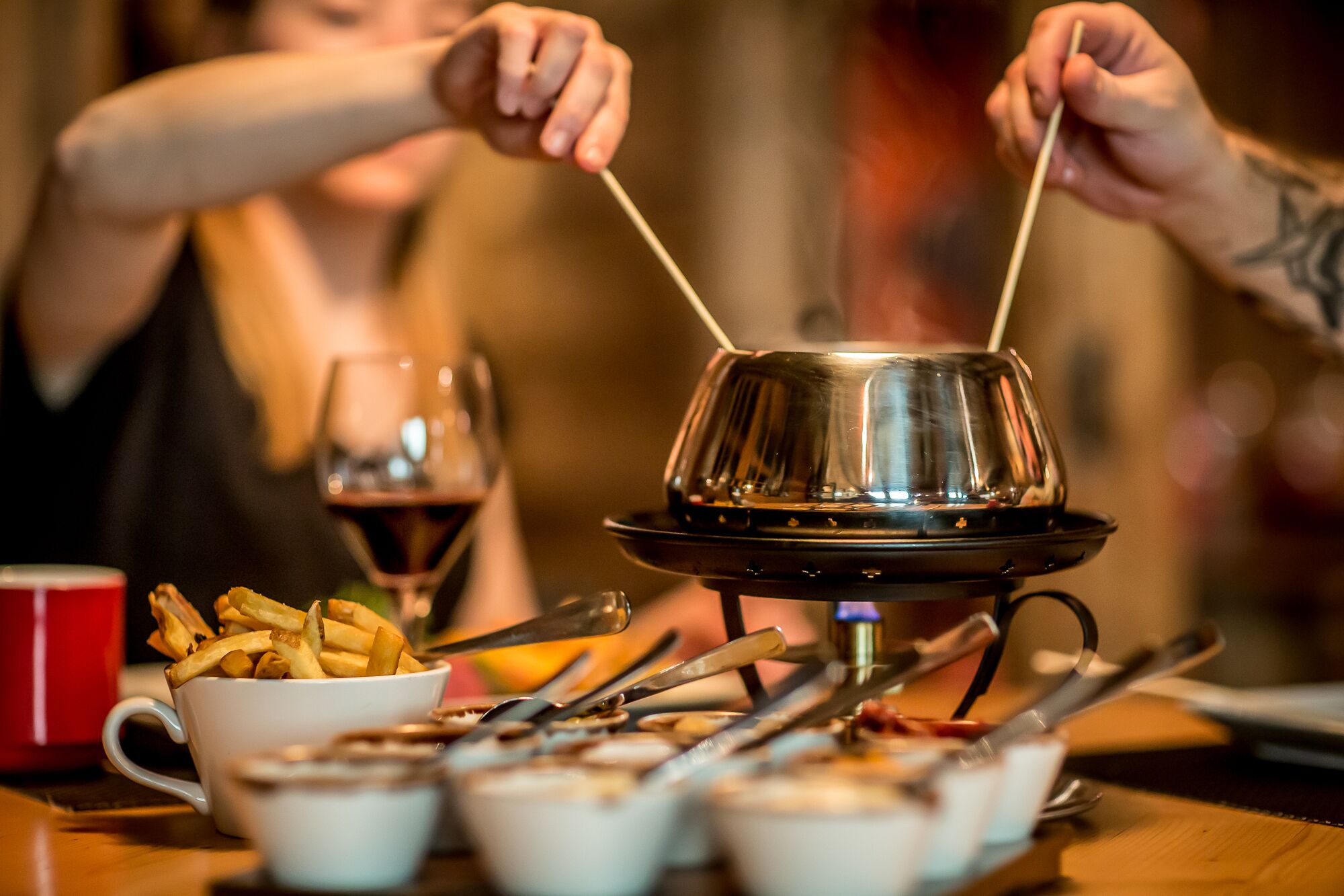 A dining table with a metal fondue pot, a bowl of fries, and a glass of red wine.