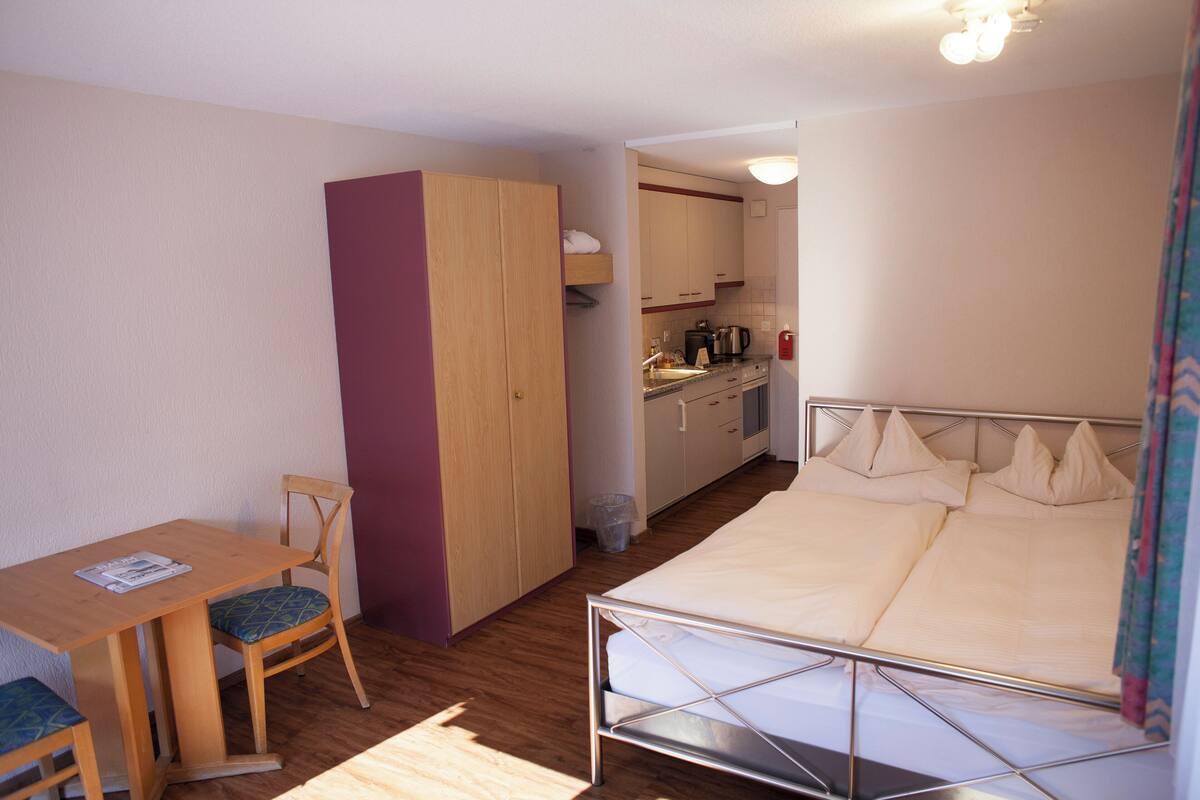 A compact hotel room with a single bed, a small desk with a chair, a kitchenette, and a wardrobe.