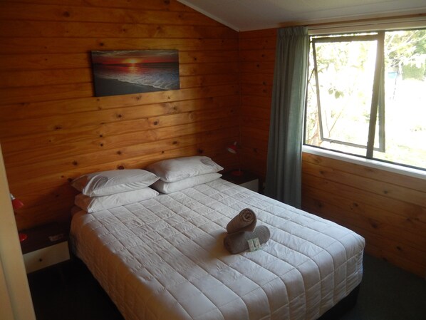 A wooden-paneled room with a bed, pillows, a nightstand, a lamp, and a painting of a sunset.