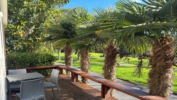 Um deck de madeira com cadeiras de metal, um banco de madeira e vista para um gramado verde e árvores.