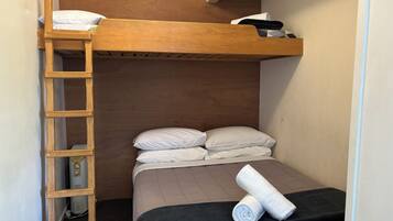 Een kleine, compacte kamer met een stapelbed, een houten trapje, een zwart matras en een opgerolde handdoek.