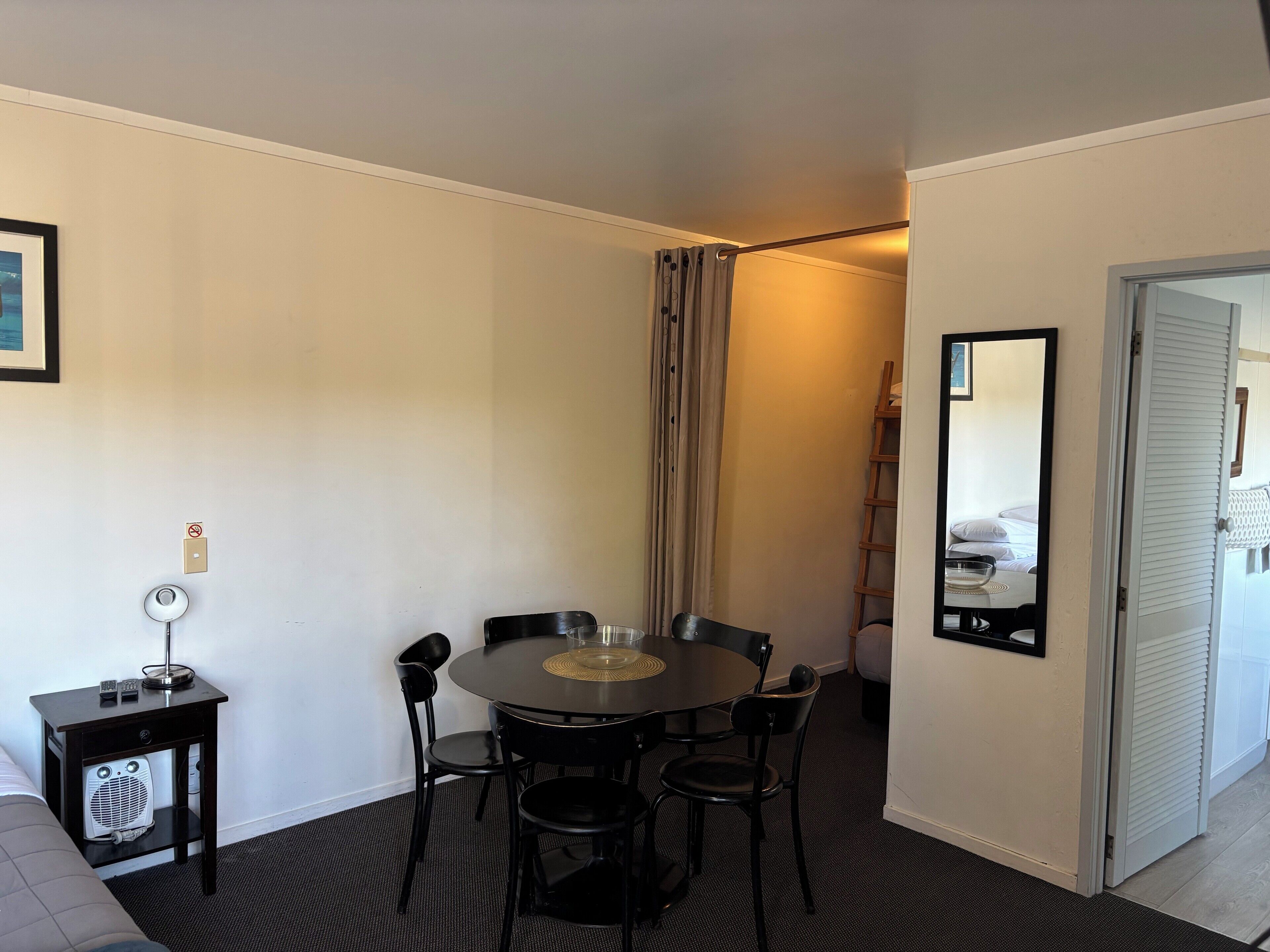 A compact hotel room with a round table and four chairs, a small bedside table with a lamp, a fan, and a mirror on the wall.