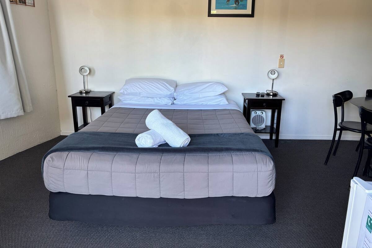 A neatly made bed with a grey quilt, two pillows, and two rolled towels. There is a framed picture on the wall above the bed.